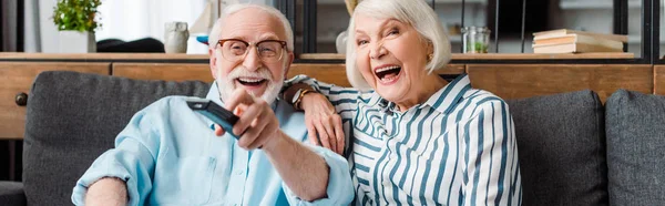 Vue panoramique du couple de personnes âgées riant tout en regardant la télévision sur le canapé à la maison — Photo de stock