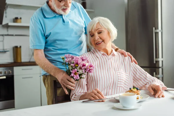 Selektiver Fokus eines älteren Mannes, der seiner lächelnden Frau während des Frühstücks in der Küche einen Strauß reicht — Stockfoto