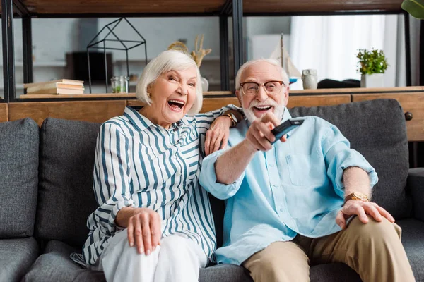 Pareja mayor riendo mientras ve la televisión en el sofá en la sala de estar - foto de stock