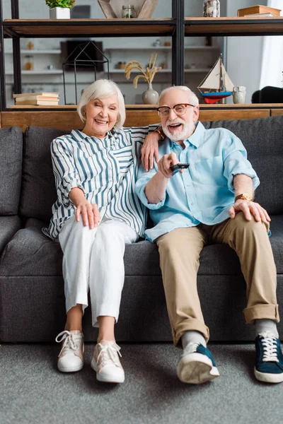 Smiling senior couple with remote controller sitting on sofa in living room — Stock Photo
