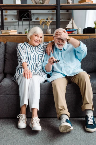 Hombre mayor con la mano por la cabeza wolding mando a distancia sonriendo esposa en el sofá - foto de stock