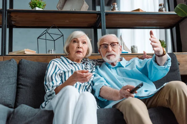 Low angle view of shocked woman watching tv by husband with remote controller on sofa — Stock Photo