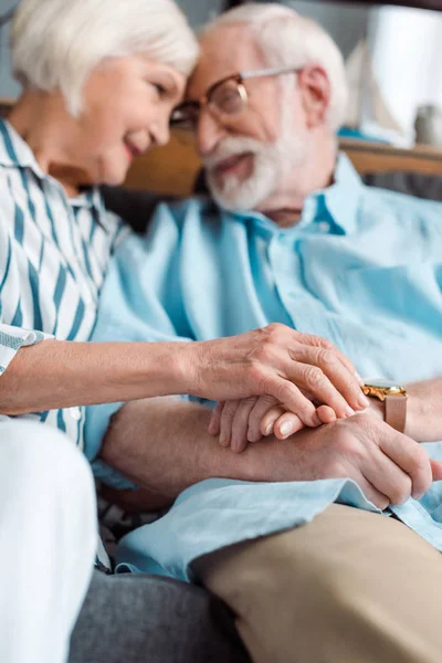 Vue à angle bas du couple de personnes âgées souriantes se tenant la main et se regardant sur le canapé — Photo de stock