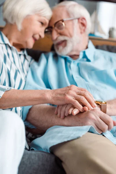 Vista de ángulo bajo de la sonriente pareja de ancianos tomados de la mano y mirándose en el sofá - foto de stock