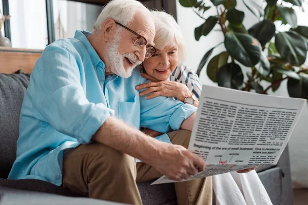Smiling senior couple reading newspaper on couch in living room — Stock Photo
