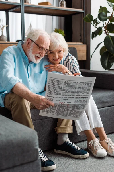 Mujer mayor sonriente sentada por el marido con el periódico en el sofá en casa - foto de stock