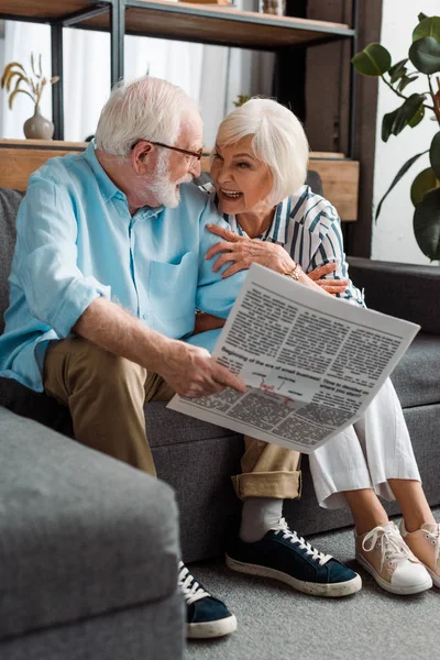 Casal sênior sorrindo um para o outro e homem segurando jornal no sofá em casa — Fotografia de Stock