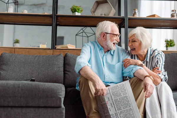 Seniorenpaar mit Zeitung lacht, während es auf Couch im Wohnzimmer sitzt — Stockfoto