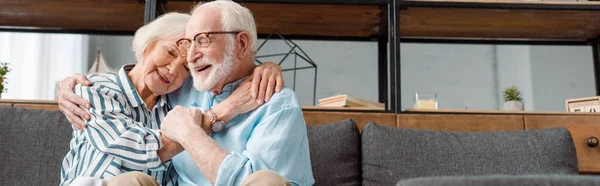 Panoramic shot of smiling senior woman hugging husband on couch at home — Stock Photo