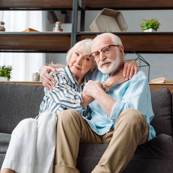 Seniorenpaar schaut in die Kamera, während es sich auf der Couch umarmt — Stockfoto