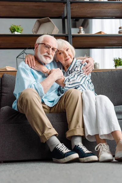 Blick auf ein älteres Paar, das sich auf dem Sofa im Wohnzimmer umarmt und in die Kamera schaut — Stockfoto