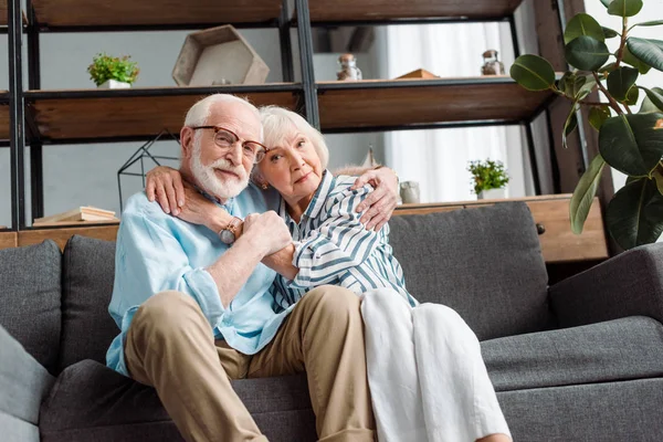 Visão de baixo ângulo da mulher idosa abraçando o marido e olhando para a câmera no sofá em casa — Fotografia de Stock