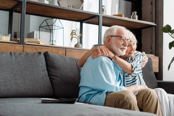 Selective focus of senior couple hugging by remote controller on couch at home — Stock Photo