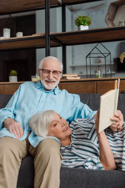 Mujer mayor leyendo libro sonriendo marido en sofá en casa - foto de stock