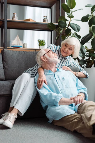 Mujer mayor sonriente abrazando marido en el suelo en la sala de estar - foto de stock