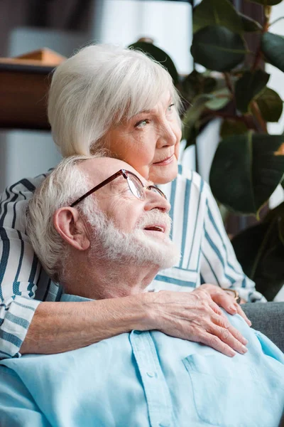 Mujer mayor abrazando marido y mirando hacia otro lado en casa - foto de stock