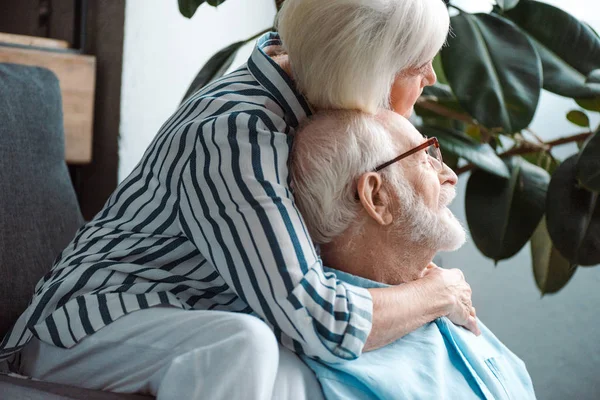Вид сбоку пожилой женщины, обнимающей мужа и отводящей взгляд в гостиной — стоковое фото