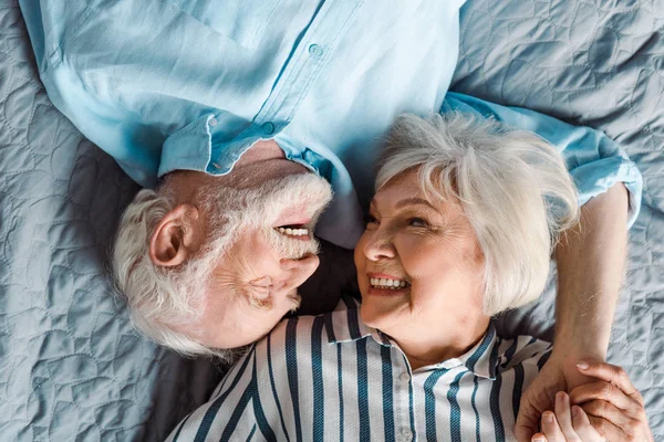 Вид сверху пожилой пары, улыбающейся друг другу и держащейся за руки на кровати — Stock Photo