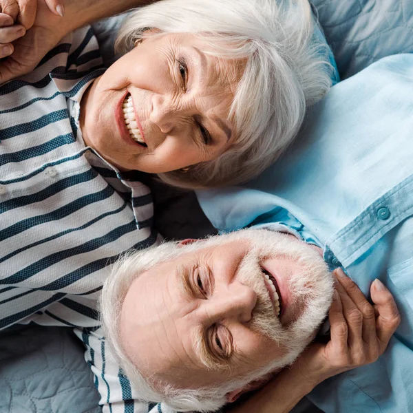 Вид сверху на пожилую пару, улыбающуюся на камеру, лежа на кровати — стоковое фото