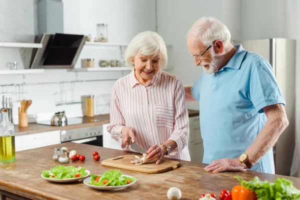 Пожилой мужчина обнимает улыбающуюся жену, режет грибы на кухонном столе — стоковое фото