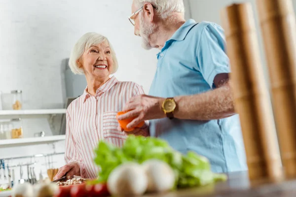 Enfoque selectivo de la mujer sonriente mirando al marido mientras cocina en la mesa de la cocina - foto de stock