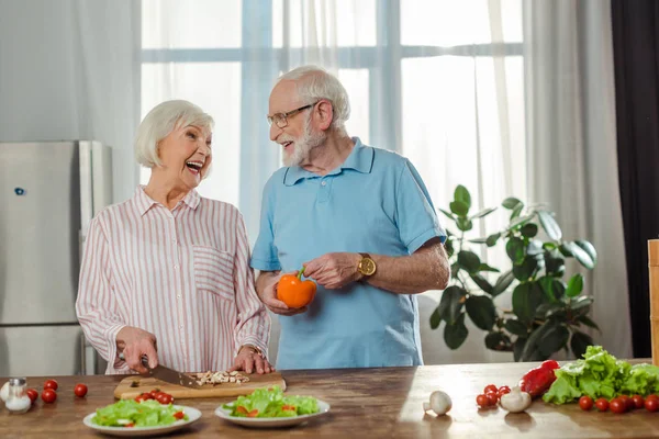 Selective focus of senior woman laughing while cutting vegetables by husband in kitchen — Stock Photo
