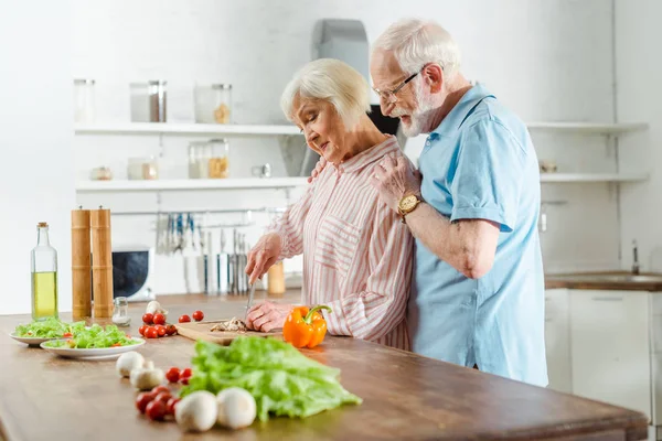 Seitenansicht eines älteren Mannes, der seine Frau beim Kochen am Küchentisch umarmt — Stockfoto