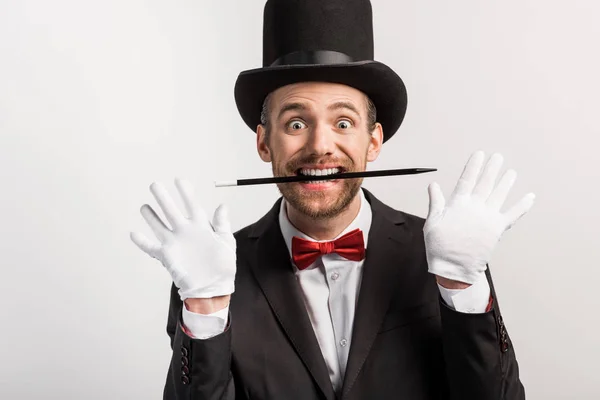 Excited magician in suit and hat holding wand in teeth, isolated on grey — Stock Photo