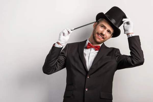 Pensativo mago sonriente en traje y sombrero sosteniendo varita, aislado en gris - foto de stock