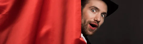 Panoramic shot of shocked handsome magician in suit and hat in circus with red curtains — Stock Photo