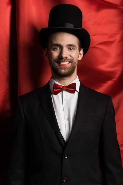 Guapo mago sonriente en traje y sombrero en circo con cortinas rojas - foto de stock