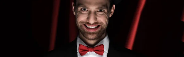 Panoramic shot of scary smiling gentleman in suit and bow tie in circus with red curtains — Stock Photo