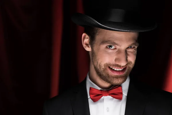 Asustadizo sonriente mago profesional en traje y sombrero en circo con cortinas rojas - foto de stock