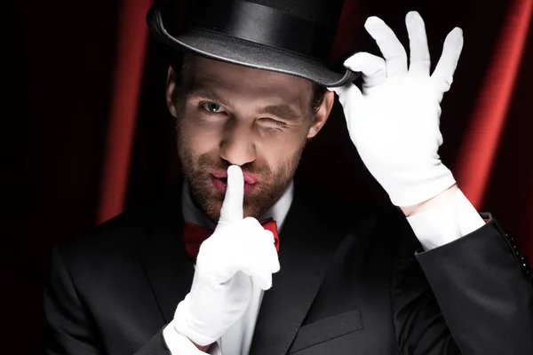 Magician winking and showing silence symbol in circus with red curtains — Stock Photo