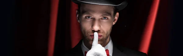 Panoramic shot of magician showing silence symbol in circus with red curtains — Stock Photo