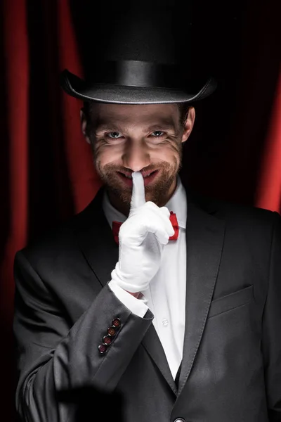 Magicien souriant montrant symbole de silence dans le cirque avec des rideaux rouges — Photo de stock