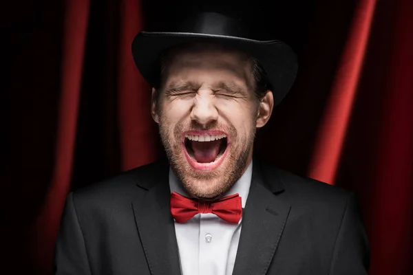 Aterrador mago emocional con los ojos cerrados y la boca abierta en circo con cortinas rojas - foto de stock