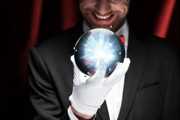 Cropped view of smiling magician holding blue magic ball in circus with red curtains — Stock Photo