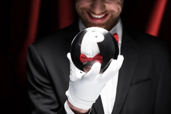 Cropped view of smiling magician holding magic ball in circus with red curtains — Stock Photo