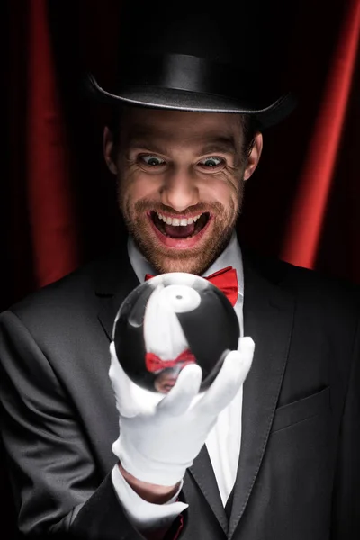 Excited scary magician looking at magic ball in circus with red curtains — Stock Photo