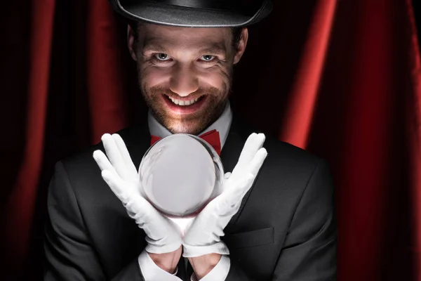 Scary smiling magician holding magic ball in circus with red curtains — Stock Photo