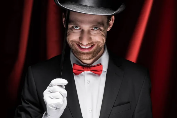 Sorrindo mágico assustador em terno e chapéu segurando varinha no circo com cortinas vermelhas — Fotografia de Stock