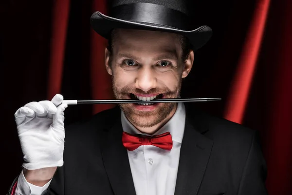 Professional scary magician in suit and hat holding wand in teeth in circus with red curtains — Stock Photo