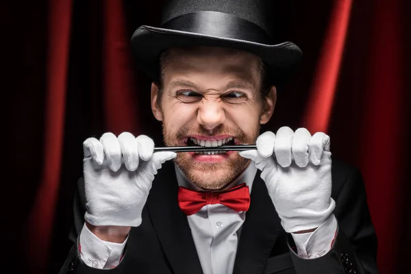 Mago asustadizo emocional en traje y sombrero sosteniendo varita en los dientes en circo con cortinas rojas - foto de stock