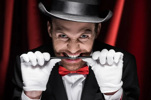 Handsome scary magician in suit and hat holding wand in teeth in circus with red curtains — Stock Photo