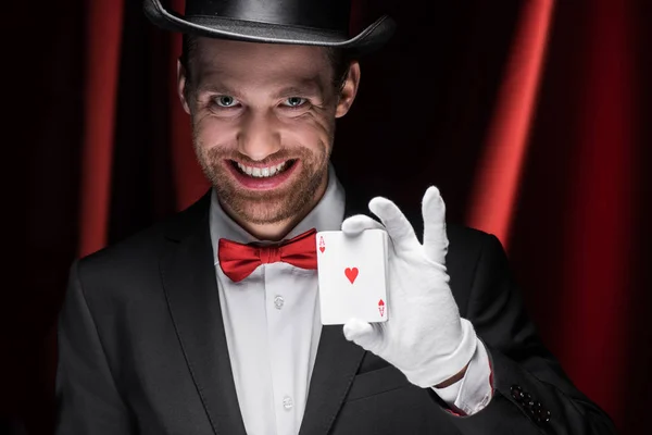 Smiling magician holding playing cards in circus with red curtains — Stock Photo