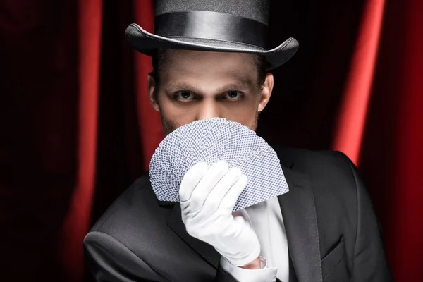 Magician in hat holding playing cards in circus with red curtains — Stock Photo