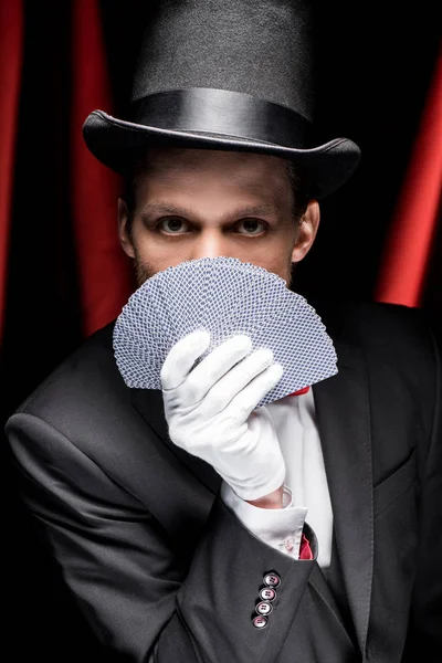 Young magician holding playing cards in circus with red curtains — Stock Photo