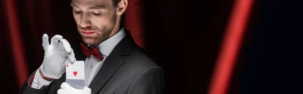 Panoramic shot of serious magician holding playing cards in circus with red curtains — Stock Photo