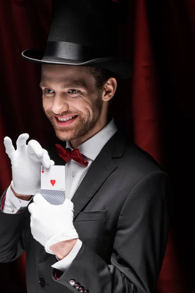 Smiling magician holding playing cards in circus with red curtains — Stock Photo